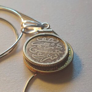 May include: A silver chain necklace with a round pendant featuring an old coin with Arabic script. The coin is surrounded by a gold-colored band.