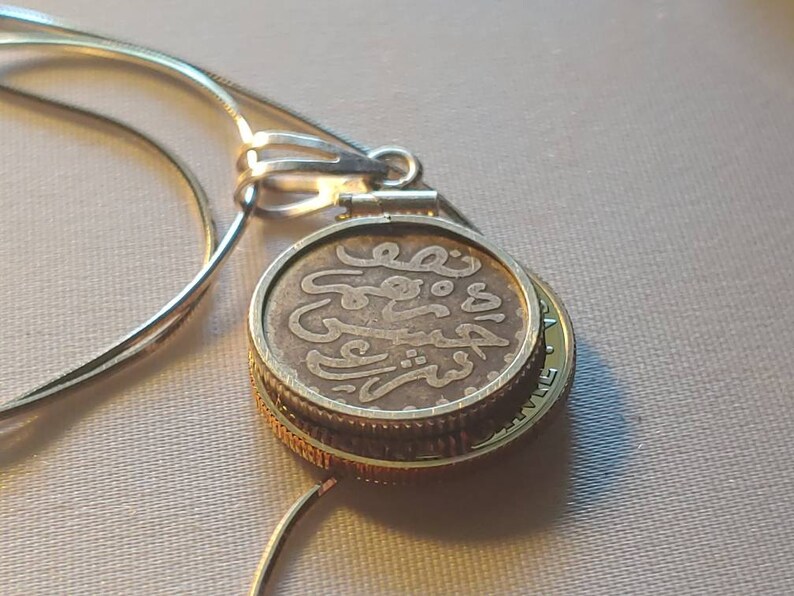 May include: A silver chain necklace with a round pendant featuring an old coin with Arabic script. The coin is surrounded by a gold-colored band.