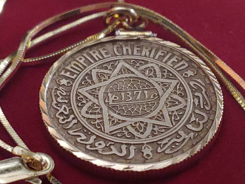 May include: A gold pendant necklace with a silver coin featuring the words "Empire Cherified" and the year "1871" in Arabic script. The coin is surrounded by a decorative border.