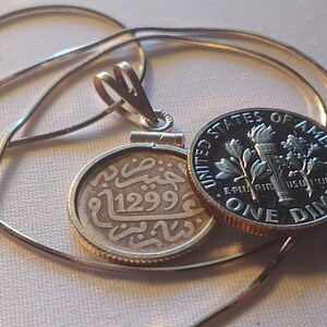 May include: A silver pendant necklace with a round coin charm featuring Arabic script and the number "1299". The pendant is attached to a silver chain and is next to a US dime.