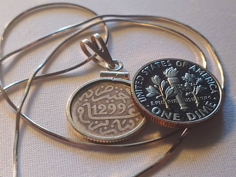 May include: A silver pendant necklace with a round coin charm featuring Arabic script and the number "1299". The pendant is attached to a silver chain and is next to a US dime.