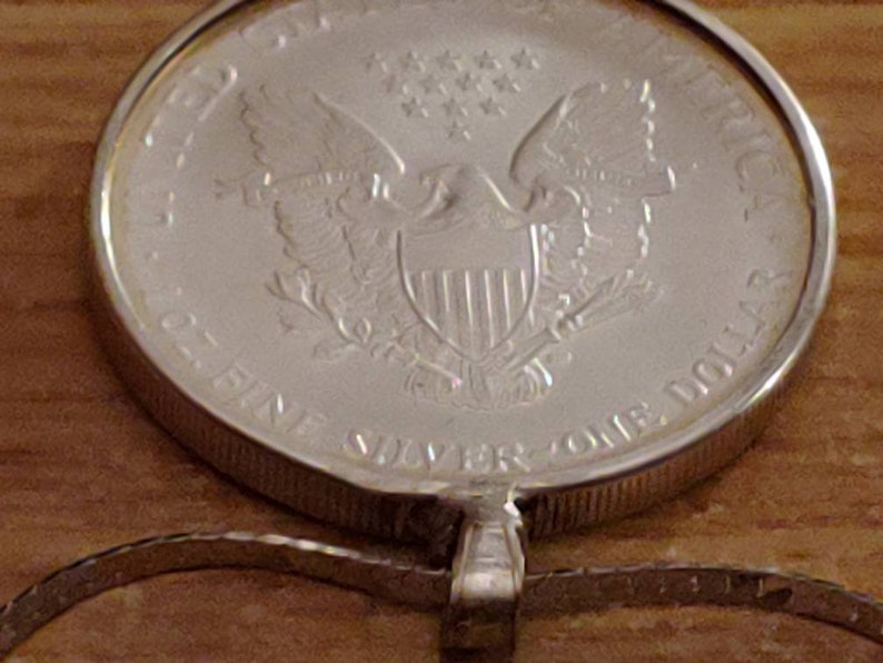 May include: A silver coin pendant featuring the American Eagle design. The coin is marked "ONE DOLLAR" and "1 OZ. FINE SILVER". The pendant is attached to a silver chain, showcasing a classic and valuable piece of jewelry.