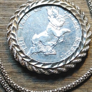 May include: Silver pendant necklace featuring a circular pendant with a raised border and a detailed image of a rabbit. The pendant is inscribed with text and hangs from a delicate silver chain. The necklace is displayed on a wooden surface.