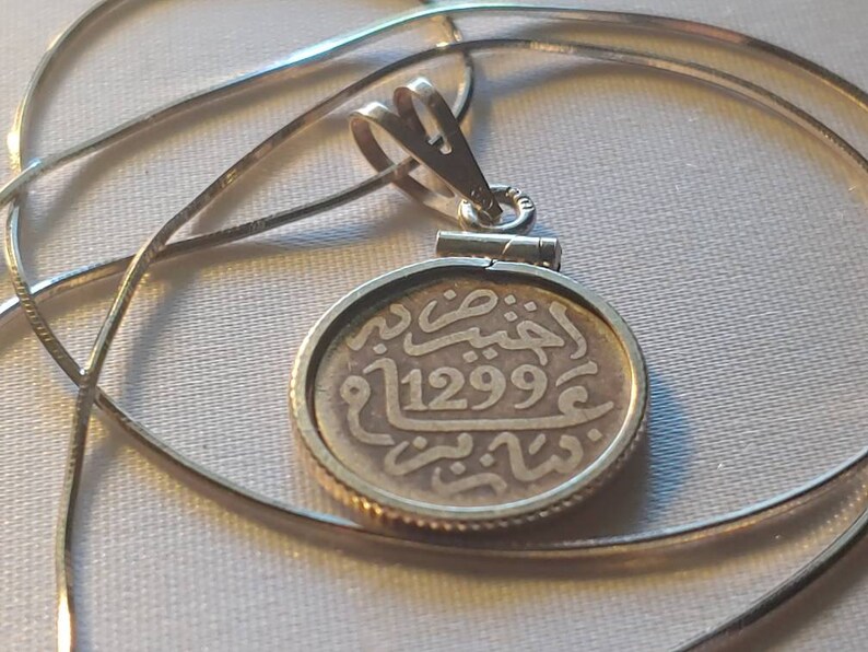 May include: A silver chain necklace with a silver pendant featuring an old coin with Arabic script and the date 1299.