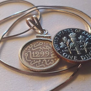 May include: A silver chain necklace with a gold pendant featuring an old coin with Arabic script and the year 1299. The coin is attached to a silver dime with the inscription "United States of America, E Pluribus Unum, One Dime".