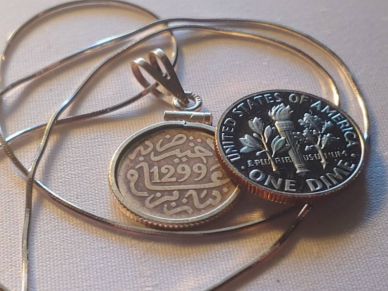 May include: A silver chain necklace with a gold pendant featuring an old coin with Arabic script and the year 1299. The coin is attached to a silver dime with the inscription "United States of America, E Pluribus Unum, One Dime".