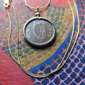 May include: A gold-colored necklace with a pendant made from an Irish coin. The coin features a harp and the year 1988. The necklace is displayed against a colorful, abstract background with red, blue, and pink hues.