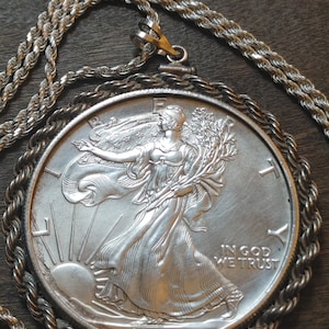 Może przedstawiać: Srebrny wisiorek z monetą American Eagle i łańcuszkiem w kształcie sznura. Na monecie widnieje wizerunek Lady Liberty idącej z pochodnią i gałązką oliwną. Monety są datowane na 1995 rok i mają napis "In God We Trust".