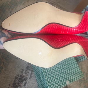 May include: A pair of bright red pointed-toe pumps featuring a textured snakeskin-like pattern on the exterior. The shoes have a slim kitten heel and a smooth, light beige sole. The vibrant red material covers the sides and back of the footwear.