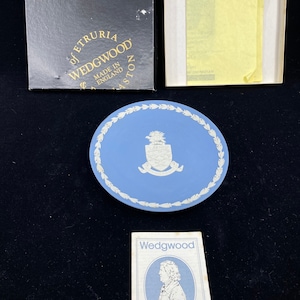 Puede incluir: Un plato de porcelana Wedgwood azul y blanco con un escudo y un borde dorado. El plato está en una caja con el logotipo de Wedgwood y las palabras "of Etruria Wedgwood Made in England" en ella. También hay una pequeña tarjeta con el logotipo de Wedgwood y las palabras "a living tradition" en ella.