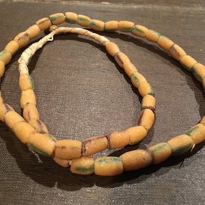 May include: A necklace of oblong beads in shades of tan, green, and purple. The beads are strung together with a natural fiber cord. The necklace is displayed against a dark background.