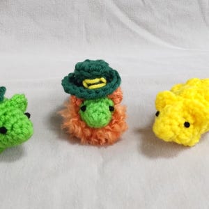 May include: Three crocheted stuffed toys. One is green with a green leaf on top, one is orange with a green hat, and one is yellow.
