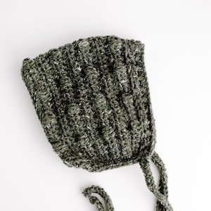 May include: A green crocheted baby bonnet with a long tie. The bonnet is made of a soft, textured yarn.