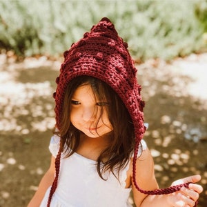 May include: A young girl wearing a maroon crocheted hooded hat with a textured bobble design. The hat has long ties that hang down the front of the girl's white dress.