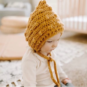 May include: A baby wearing a crocheted bonnet with a textured pattern in a mustard yellow color. The bonnet has a long tie that hangs down the front of the baby's chest.