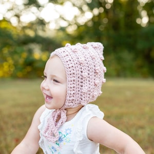 May include: A young child wearing a pink crocheted bonnet with a white dress and floral embroidery. The child is smiling and running through a grassy field.