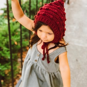 May include: A deep burgundy crochet bonnet featuring a textured bobble stitch pattern and a pointed top. The bonnet includes two matching crocheted ties that fasten under the chin, providing a secure fit with a distinct, handcrafted appearance.
