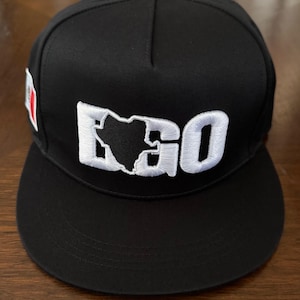 May include: Black baseball cap with the word "EGO" embroidered in white. The "O" features a white outline of a country's shape. A small flag is on the side of the cap.