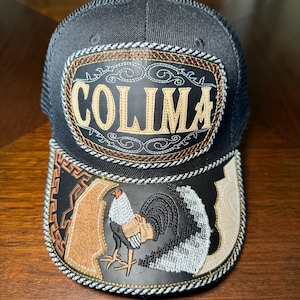May include: A black trucker hat featuring a central patch with the text COLIMA in gold embroidery. The brim displays a detailed embroidered rooster graphic, geometric patterns, and decorative stitching in shades of black, tan, and white.