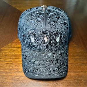May include: A black baseball cap featuring an all-over embroidered skull pattern. The front panel displays three large, black, beaded letters with a central hand gesture icon. A small, metallic, cross-shaped button sits at the top center of the cap.