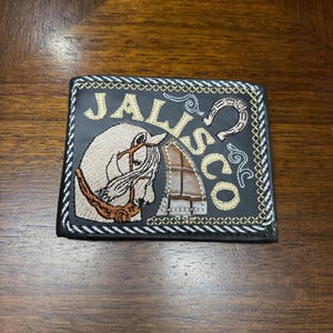 Puede incluir: Cartera de cuero negro con un caballo blanco y la palabra "JALISCO" bordada en hilo dorado. La cartera tiene un borde negro y blanco.