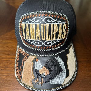 May include: A black baseball cap featuring a rectangular patch with the word TAMAULIPAS embroidered in gold lettering. The brim displays a detailed embroidered rooster graphic, geometric patterns in tan and black, and decorative silver rope trim.