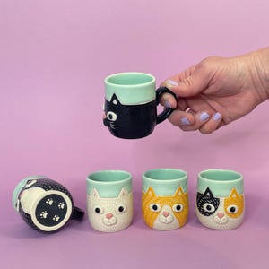 May include: A set of five ceramic mugs featuring cat faces with raised details. Each mug has a light mint green rim and a unique cat design, including black, white, orange tabby, and calico patterns. One mug shows a paw print on the base.