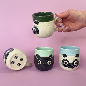 May include: A collection of ceramic mugs featuring pug dog designs. The mugs have a variety of colors, including white, black, and blue, with green and blue rims. One mug is held by a hand. The bottom of one mug has paw prints.