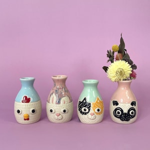 May include: Four ceramic vases with animal faces: a chicken, a rabbit, a cat, and a pug. The chicken vase is light blue and white, the rabbit vase is pink and white, the cat vase is mint green and white, and the pug vase is pink and black. The pug vase holds flowers.