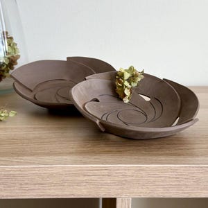 May include: Two brown ceramic bowls with a unique, swirling design. Each bowl has a petal-like shape and a decorative cutout. Dried flowers are placed in and around the bowls, adding a natural element to the composition.