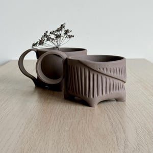 Peut inclure: Deux tasses en céramique marron avec des motifs texturés uniques. L'une a une anse arrondie, l'autre présente un motif vertical sculpté. Une brindille de fleurs séchées est dans une tasse. Les tasses sont sur une surface en bois clair.