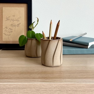 以下が含まれることがあります： テクスチャードデザインのベージュ色のセラミックペンシルホルダー。3本の鉛筆が入っています。小さな鉢植えの植物がペンシルホルダーの隣にあります。額入りのアート作品とノートの束が背景にあります。