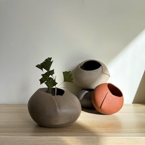 Könnte beinhalten: Eine Sammlung moderner Keramikvasen in verschiedenen Erdtönen. Eine Vase enthält einen Zweig mit Grünzeug. Die Vasen sind kugelförmig mit einer einzigartigen, asymmetrischen Öffnung. Die Farbpalette umfasst Braun-, Grau- und Terrakotta-Töne.