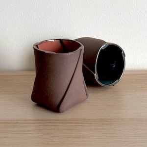Puede incluir: Dos tazas de cerámica marrón con un diseño plegado único. Las tazas tienen un acabado mate y un interior marrón rojizo. Una taza está en posición vertical, la otra de lado, revelando un borde metálico.
