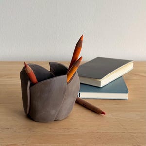 May include: A gray ceramic pencil holder shaped like a flower, holding several orange and brown pencils. Two notebooks are stacked in the background. The holder sits on a wooden surface.