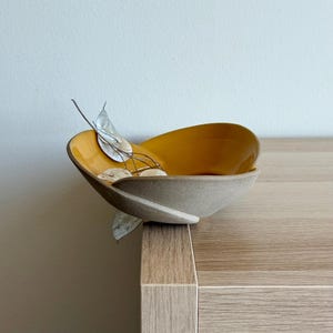 Może przedstawiać: Dekoracyjna ceramiczna miska z musztardowym wnętrzem i szarym zewnętrzem. Misa zawiera małe, jasne przedmioty i kilka suchych łodyg. Misa stoi na jasnej drewnianej powierzchni.