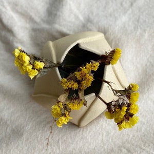Könnte beinhalten: Eine weiße Keramikvase mit geometrischem Design, gefüllt mit getrockneten gelben Blumen.