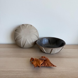 May include: A small, beige ceramic bowl with a dark interior and a matching lid. The lid has a textured, floral-like design. A few dried leaves are in front of the bowl, on a light wood surface.