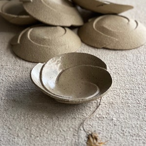 Puede incluir: Un juego de pequeños cuencos de cerámica beige con un diseño de textura en espiral. Los cuencos están apilados uno encima del otro, y un cuenco está sentado sobre una superficie de tela beige.