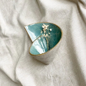 Può includere: Una ciotola in ceramica blu-verde con un bordo dorato, a forma di conchiglia, con fiori bianchi secchi all'interno.