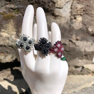 May include: Three macrame rings with different coloured cords and beads. The rings are in the shape of a four-leaf clover. The beads are black, grey, and pink.
