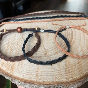 May include: Four braided leather bracelets in brown, black, and tan. The bracelets are on a wooden surface.