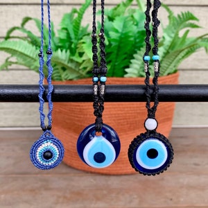 May include: Three evil eye necklaces with blue and black glass beads and a black cord. The necklaces are hanging from a black bar.