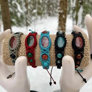 Peut inclure: Cinq bracelets en macramé avec des cordons de différentes couleurs et des perles de pierres précieuses. Les bracelets sont présentés sur un rouleau de toile de jute brune.