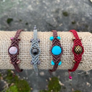 Peut inclure: Quatre bracelets en macramé faits à la main avec des cordons et des perles de différentes couleurs. Les bracelets présentent une pierre turquoise, une pierre brune, un charme en métal et un bouton avec un motif.