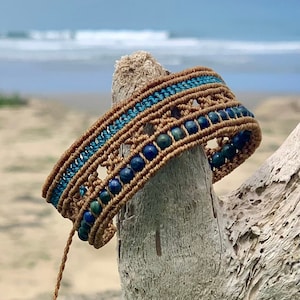 May include: A brown and blue beaded bracelet with a woven design. The bracelet is made with small, round beads and is tied around a piece of driftwood.