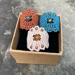 May include: Three macrame rings with different coloured cords and gemstone accents. The rings are displayed in a brown box with a black interior.