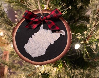 May include: A wooden Christmas ornament shaped like the state of West Virginia, painted white on a black background. The ornament is decorated with a red and black plaid bow.