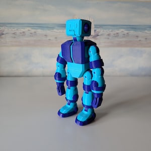 Puede incluir: Un juguete robot articulado azul y morado con una cabeza cuadrada. El robot tiene un cuerpo azul con detalles morados y está en posición de pie con los brazos a los lados.