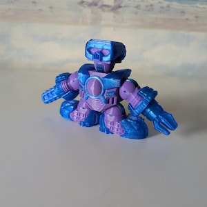 Puede incluir: Una figura de robot azul y morado con una cabeza cuadrada, hombros anchos y manos en forma de garra. El robot de juguete tiene un diseño circular en el pecho y está en posición sentada. El juguete es de plástico.